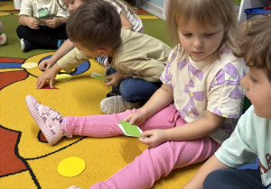 Dzieci siedzą na dywanie a w rączkach trzymają wycięte figury geometryczne