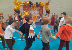 Dzieci tańczą w dużym kole trzymając się za ręce. W tle baner z motywem jesiennym i balonowa girlanda.