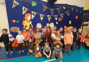 Dzieci w jesiennych strojach pozują do wspólnego zdjęcia na tle dekoracji z napisem Bal Jesienny.