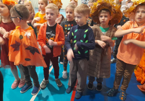 Dzieci w jesiennych strojach stoją obok siebie i wykonują młynek rękami przy piosence.