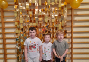 Trzech chłopców pozuje do wspólnego zdjęcia na tle dekoracji - na drabinkach wiszą złote paski, a na górze balonowa girlanda.