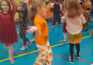 Na pierwszym planie grupa dziewczynek tańczy w małym kółeczku z rękami podniesionymi do góry. W tle inni uczestnicy balu.