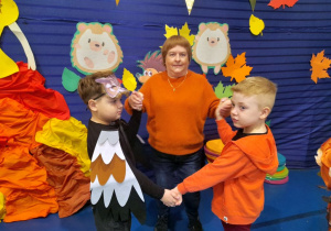 Dwóch chłopców tańczy z Panią Dorotką w kółeczku. W tle jesienna dekoracja.