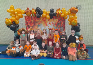 Dzieci w jesiennych strojach pozują do wspólnego zdjęcia. W tle baner z motywem jesiennym i balonowa girlanda.