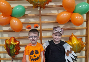 Mikołaj i Julek w jesiennych strojach stoją obok siebie na tle drabinek ozdobionych balonami.