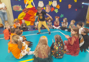 Dzieci siedzą w kole na podłodze, a Marcel w środku tańczy z kółkiem.