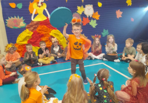 Dzieci siedzą w kole na podłodze i klaszczą w ręce, a Mikołaj tańczy z kółkiem w środku koła przy muzyce.