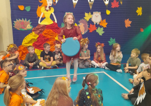 Dzieci siedzą w kole na podłodze, a Weronika tańczy z kółkiem w środku koła przy muzyce. W tle dekoracja z napisem Bal Jesienny.