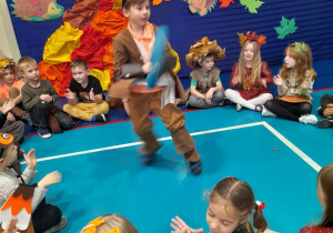 "Słoneczka" siedzą na podłodze i klaszczą w ręce, a Staś tańczy z kółkiem w środku koła przy muzyce. W tle jesienna dekoracja.