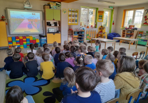 Dzieci siedzą na dywanie i oglądają film pt. "Podróże z Ambasadorką Bezpieczeństwa".