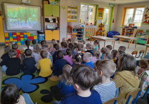 Dzieci ze wszystkich grup siedzą na dywanie i oglądają kolejny fragment filmu pt. "Podróże z Ambasadorką Bezpieczeństwa".