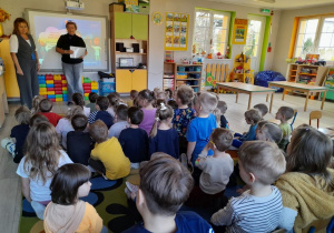 Dzieci siedzą na dywanie, a przed nimi stoją dwie przedstawicielki PKP Polskie Linie Kolejowe S.A. Panie rozmawiają z dziećmi o zasadach bezpieczeństwa podczas przekraczania przejazdów kolejowo-drogowych.