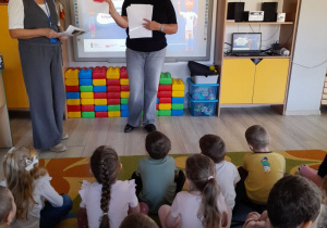 Przedszkolaki siedzą na dywanie, a przed nimi stoją dwie przedstawicielki PKP Polskie Linie Kolejowe S.A. Pani pokazuje dzieciom znak Stop.