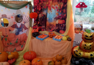 Na tablicy znajduje się jesienny baner, na szafce okrytej pomarańczowym materiałem leżą dynie, upominki dla dzieci oraz cukierki. Z obu stron tablicy przypięty jest żółto-pomarańczowy materiał oraz dyniowa fotobudka. Na podłodze leżą dynie oraz balonowe dekoracje dyniowe.