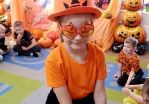 Marysia częstuje dzieci siedzące na dywanie cukierkami. Dziewczynka ma założony dyniowy kapelusz i dyniowe okulary.