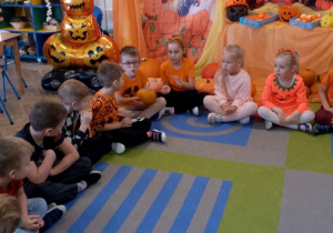 Dzieci siedzą w kole na dywanie i podają sobie małą dynię podczas zabawy "Dynia parzy". W tle dekoracja z okazji Święta Dyni.