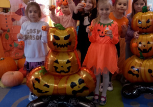 Grupka dziewczynek pozuje do zdjęcia na tle dekoracji z okazji Dnia Dyni z dyniowymi balonami i otrzymanymi upominkami.