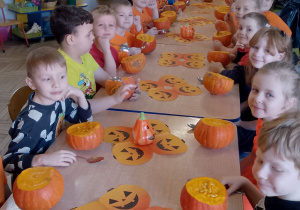 Dzieci siedzą przy wspólnym stole, na którym znajdują się dyniowe serwetki i dynie.