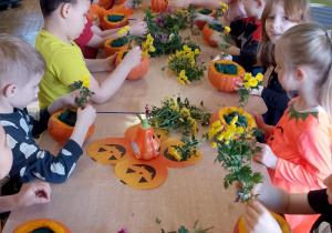 Dzieci siedzą przy wspólnym stole i dekorują dynie jesiennymi kwiatami.