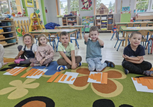 Maja, Oliwka, Oskar, Marcel i Szymek siedzą na dywanie. Przed dziećmi na białych kartonach leża ułożone w ciągu rosnącym pomarańczowe prostokąty.