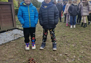 Wojtuś i Lucjan stoją w ogrodzie przedszkolnym. Przed chłopcami widać ułożoną z kasztanów strzałkę.