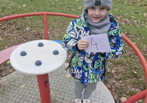 Oluś stoi wewnątrz przedszkolnej karuzeli. Chłopiec pokazuje kopertę z cyfrą 4.