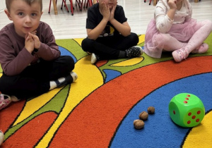 Dzieci siedzą na dywanie i przeliczają ilość kropek na kostce oraz ilość orzechów włoskich.