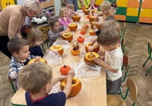 Dzieci siedzą przy stoliczkach oraz wydrążają środek dyni
