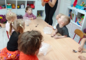 Pani Kasia prezentuje dzieciom dynię, maluchy siedzą wzdłuż stołu i uważnie słuchają Pani Kasi.