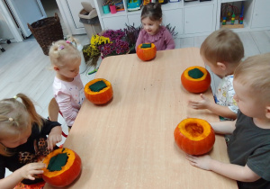 Pszczółki siedzą wzdłuż stołu, przed nimi wydrążone dynie, w których jest gąbka florystyczna.