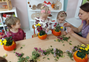 Laura, Ala i Marysia układają kompozycje z żółtych i bordowych kwiatów w dyniowej donicy, obok Pani Kasia.