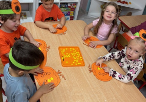 Dzieci siedzą przy stoliczkach i wyklejają plasteliną pestki w wyciętej dyni.