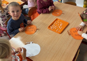Dzieci siedzą przy stoliczkach i wyklejają pestki w wyciętej dyni.