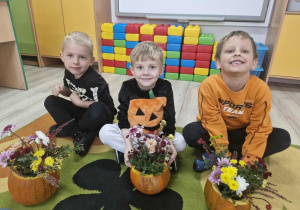 Lucjan, Oluś i Franuś siedzą na dywanie. Przed chłopcami leża skończone jesienne kompozycje z kwiatów i dyni