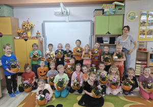 Przedszkolaki pozują do zdjęcia z Panią Kasią. Dzieci trzymają w dłoniach swoje dyniowe doniczki pełne jesiennych kwiatów