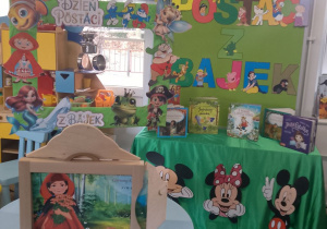 Dekoracja z okazji Dnia Postaci z Bajek: na zielonejtablicy przypięty jest kolorowy napis Dzień Postaci z Bajek oraz różne bajkowe postacie obok wisi bajkowa fotobudka. Przed tablicą na stoliku stoi teatrzyk Kamishibai z bajką "Czerwony kapturek".
