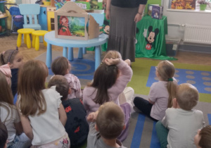 Dzieci siedzą na dywanie, przed nimi stoi pani z biblioteki. W tle dekoracja z okazji Dnia Postaci z Bajek.