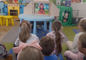 Przedszkolaki siedzą na dywanie i oglądają bajkę pt. "Czerwony kapturek" w Teatrzyku Kamishibai.