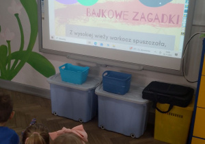 Dzieci siedzą na dywanie przed tablicą interaktywną, na której znajdują się "Bajkowe zagadki".