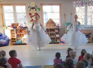 Dzieci siedzą na dywanie, a przed nimi dwie tancerki prezentują białe suknie. W tle okna, kąciki zainteresowań.