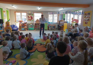 Dzieci siedzą na dywanie, a przed nimi stoi Pan i wita wszystkich na przedstawieniu baletowym. W tle okna i kąciki zainteresowań.