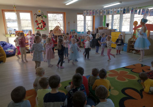 Chłopcy siedzą na dywanie, a przed nimi dziewczynki tańczą "Taniec lalek" naśladując tancerkę.