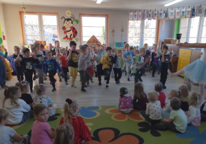 Dziewczynki siedzą na dywanie, a przed nimi chłopcy tańczą "Taniec robotów" razem z tancerką.