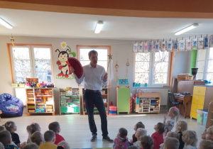 Pan stoi przed dziećmi i prezentuje rekwizyty - wachlarz oraz różdżkę. W tle okna i kąciki zainteresowań.