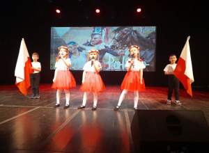 Zespół "Stokrotki" śpiewa na scenie refren piosenki "Symbole narodowe". W tle dekoracja patriotyczna.