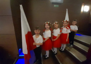 Dzieci w oczekiwaniu na występ stoją jedno za drugim na schodach w sali widowiskowej Kutnowskiego Domu Kultury. Dwóch chłopców trzyma flagi biało-czerwone. Trzy dziewczynki ubrane są w czerwone spódnice i białe koszulki, a na głowach mają biało-czerwone wianki.