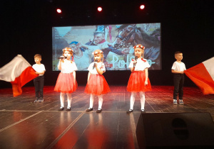 Pięcioro dzieci stoi na scenie podczas występu. Dziewczynki ubrane są w czerwone spódnice i białe koszulki, a na głowach mają biało-czerwone wianki. Chłopcy stoją po bokach trzymając biało-czerwone flagi. W tle dekoracja patriotyczna.