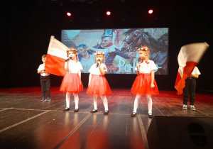 Dzieci występują na scenie w Kutnowskim Domu Kultury. Dziewczynki śpiewają piosenkę "Symbole narodowe" trzymając mikrofony w rękach. Chłopcy stoją po bokach i poruszają biało-czerwonymi flagami.