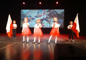 Zespół "Stokrotki" śpiewa na scenie refren piosenki "Symbole narodowe". W tle dekoracja patriotyczna.