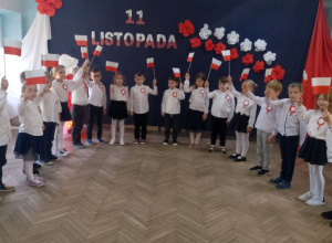 Dzieci z grupy "Słoneczka" stoją ubrane w stroje galowe z przypiętymi kotylionami w barwach narodowych. W rękach trzymają biało-czerwone flagi. W tle dekoracja z okazji 11 listopada.